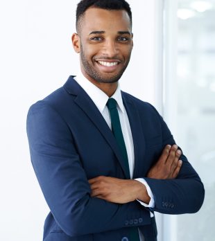 A handsome young african american businessman standing indoors with his arms folded.